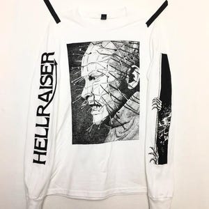 Peut inclure: T-shirt blanc à manches longues avec un graphisme noir et blanc d'un personnage avec des épingles dans la tête. Le mot "HELLRAISER" est imprimé verticalement sur la manche gauche. Un graphisme noir et blanc est sur la manche droite.
