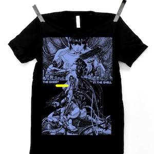 May include: Black t-shirt featuring a blue and white graphic from "Ghost in the Shell." The design includes two figures, futuristic cityscapes, and the text "THE GHOST IN THE SHELL."