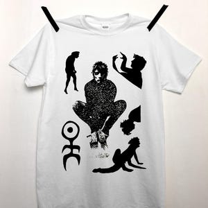 以下が含まれることがあります： 黒いグラフィックデザインの白いTシャツ。デザインは、さまざまなポーズのいくつかの人間の姿と抽象的なシンボルを特徴としています。シャツは黒いクリップが付いた物干しロープから吊り下げられています。