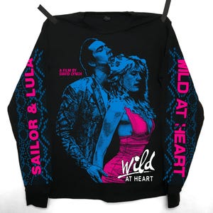 Puede incluir: Camiseta negra de manga larga con un estampado gráfico de una pareja de la película "Wild at Heart: A Film by David Lynch". Las mangas tienen un patrón de piel de serpiente con el texto "Sailor & Lula" en la manga izquierda y "Wild at Heart" en la manga derecha.