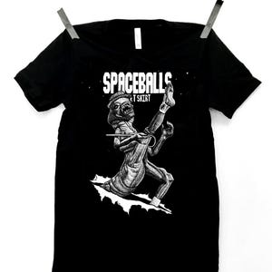 May include: Black t-shirt with the text "SPACEBALLS T SHIRT" in white. The shirt features a black and white illustration of a creature from the movie Spaceballs. The shirt is hanging from two silver clips.