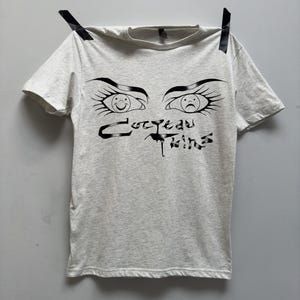May include: A light grey t-shirt with a black graphic of two eyes with smiley and sad faces. The text "CUTE SAD THINGS" is printed below the eyes.