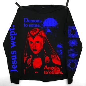 May include: Black long-sleeve shirt with a red and blue graphic design. The design features a portrait of a woman, the text "Demons to some. Angels to others." and the words "Jesus wept." on the sleeve.