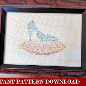 Op de afbeelding: Een kruissteekpatroon van een blauwe glazen slipper op een rood en geel kussen. Het patroon is ingelijst in een donkerhouten lijst.