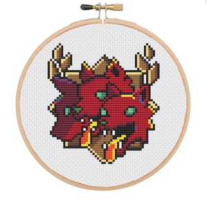 May include: A cross-stitch embroidery hoop with a pixelated design of three red dragon heads with green eyes and yellow accents. The dragons are set against a white background, framed by a wooden hoop.