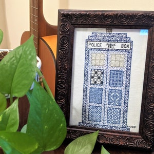 May include: A framed cross-stitch of a blue police box with the words "Police Public Call Box" on the front. The cross-stitch is in a brown frame with ornate detailing.