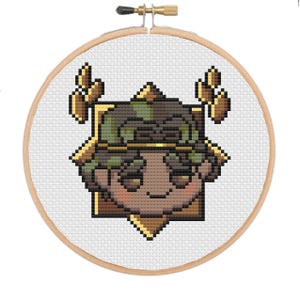 Op de afbeelding: Een kruissteekborduurwerk in een houten hoepel. Het ontwerp toont een gepixeld personage met een bruin gezicht, groene en bruine camouflage-hoofddeksels en gouden accenten. De achtergrond is wit.