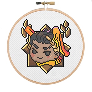 May include: A cross-stitch embroidery hoop featuring a pixelated portrait of a person with brown skin, dark hair, and colorful accents. The design includes shades of brown, orange, yellow, and pink, set against a white background. The wooden hoop is visible.