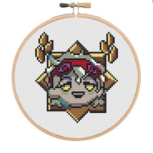 以下が含まれることがあります： 星型のフレームの中に、様式化されたキャラクターのクロスステッチ刺繍。キャラクターは花の冠を持ち、茶色、グレー、赤、黄色のカラーパレットを使用しています。刺繍は木製のフレームにセットされています。