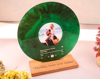 Puede incluir: Un disco de vinilo verde con una foto de un padre y un hijo en la parte delantera. El disco está en un soporte de madera con el texto "Daddy, love you more" grabado en él. El disco está reproduciendo la canción "Runnin' Down A Dream" de Tom Petty.