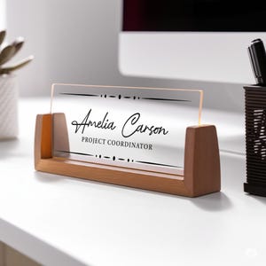 May include: A wooden desk nameplate with a clear acrylic panel. The name "Amelia Carson" and the title "Project Coordinator" are printed in black. The nameplate has a light brown wooden base and supports.