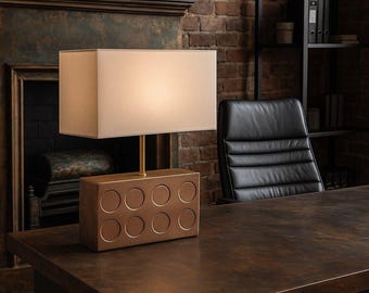 Könnte beinhalten: Eine braune Tischlampe mit einem weißen Stoffschirm. Der Lampenfuß ist in Form eines großen Lego-Steins mit einem goldenen Metallstiel geformt. Die Lampe steht auf einem dunkelbraunen Holztisch.