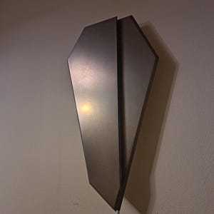 May include: A contemporary wall light with a metallic finish. The light features a distinctive, angular design with two overlapping panels. A white cable extends from the bottom. The light is mounted on a light-coloured wall.