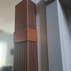 May include: A copper-coloured wall sconce with a rectangular shape and vertical lines. The sconce is mounted on a grey wall.