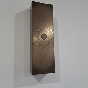 May include: A rectangular wall sculpture with a bronze finish. The sculpture features a raised, circular design in the centre with a textured surface. The design resembles a sun with radiating lines.