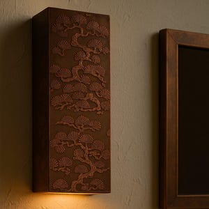 May include: A bronze-coloured wall sconce with a detailed relief of a stylized tree design. The sconce is mounted on a textured wall, with a warm light emanating from the bottom. A framed mirror is to the right.