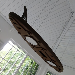 May include: A brown surfboard-shaped light fixture with a fin and a star emblem, suspended from the ceiling. The fixture has two circular light fixtures and is mounted in a room with large windows.
