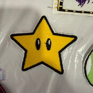 May include: Embroidered yellow star patch with a black outline and two oval eyes. The star is a five-pointed shape, with a textured surface. The patch is likely for clothing or accessories.