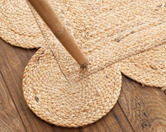 May include: A light tan, braided jute rug featuring a scalloped edge design with rounded protrusions. A smooth, cylindrical wooden furniture leg rests on the textured surface of the rug, which displays a consistent woven pattern throughout.