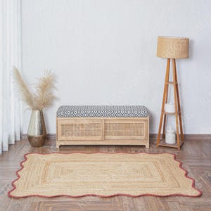 Puede incluir: Una alfombra rectangular de yute con borde festoneado y un fino ribete rojo. La alfombra presenta una textura tejida en tono bronceado natural. Se sit&uacute;a sobre un suelo de madera frente a un banco de almacenamiento de madera clara con rejilla.