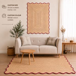 Puede incluir: Una alfombra rectangular de yute trenzado con un borde festoneado acabado en un ribete rojo oscuro. La alfombra se muestra tanto en el suelo como colgada en la pared. El texto indica que hay tama&ntilde;os y colores personalizados disponibles.