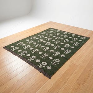 Könnte beinhalten: Ein rechteckiger, grüner Teppich mit einem sich wiederholenden weißen Blumenmuster. Der Teppich hat einen Fransenrand und wird auf einem Holzboden präsentiert. Der Teppich ist ein dekoratives Wohnaccessoire.
