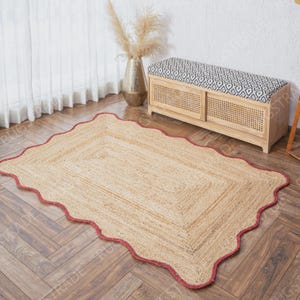 Puede incluir: Una alfombra rectangular hecha de fibras trenzadas de color tostado natural con un borde festoneado rematado por una fina l&iacute;nea roja. La alfombra tiene una superficie plana y texturizada con un patr&oacute;n rectangular conc&eacute;ntrico en el centro.