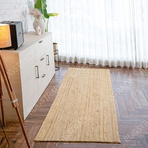 Puede incluir: Una alfombra rectangular de yute beige con un diseño texturizado y acanalado. La alfombra está colocada sobre un suelo de madera oscura, frente a un mueble blanco y una ventana con cortinas blancas. Una lámpara de pie es visible a la izquierda.