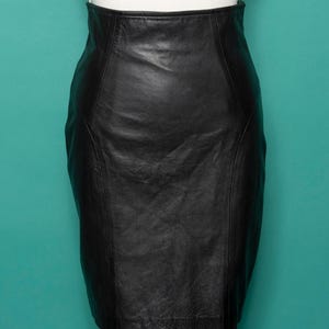 May include: A black leather pencil skirt with a high waist. The skirt is displayed on a mannequin against a teal background. The skirt has a classic, fitted silhouette.