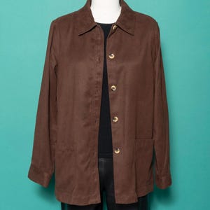 May include: A brown button-up jacket with a collared neckline and two front pockets. The jacket has long sleeves and gold-colored buttons. The jacket is displayed on a mannequin against a teal background. The jacket is made of a suede-like material.