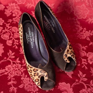 K&ouml;nnte beinhalten: Ein Paar Peep-Toe-Pumps aus dunkelbraunem Wildleder mit Leopardenmuster-Akzenten. Die Innensohlen sind mit dem Text COUTURE DONALD J PLINER MADE IN THE MOUNTAINS OF ITALY versehen. Die Schuhe haben einen hohen Absatz und ein geschwungenes Design.