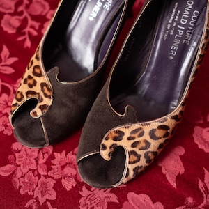 K&ouml;nnte beinhalten: Ein Paar Donald J Pliner Couture Peeptoe-Pumps mit einer Vorderseite aus dunkelbraunem Wildleder und einem Leopardenmuster-Overlay aus Kalbshaar. Das Innenfutter ist dunkelviolett mit dem Markennamen und Made in the Mountains of Italy in Wei&szlig;.
