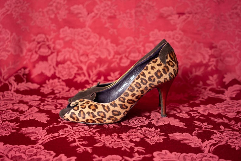 K&ouml;nnte beinhalten: Ein Paar High-Heel-Pumps mit offener Zehenpartie und einem beigefarbenen und schwarzen Leopardenmuster. Die Schuhe haben einen dunkelbraunen Besatz am Kragen und an der Ferse, ein dunkles Innenfutter, einen schlanken Absatz und ein gewelltes Randdetail.