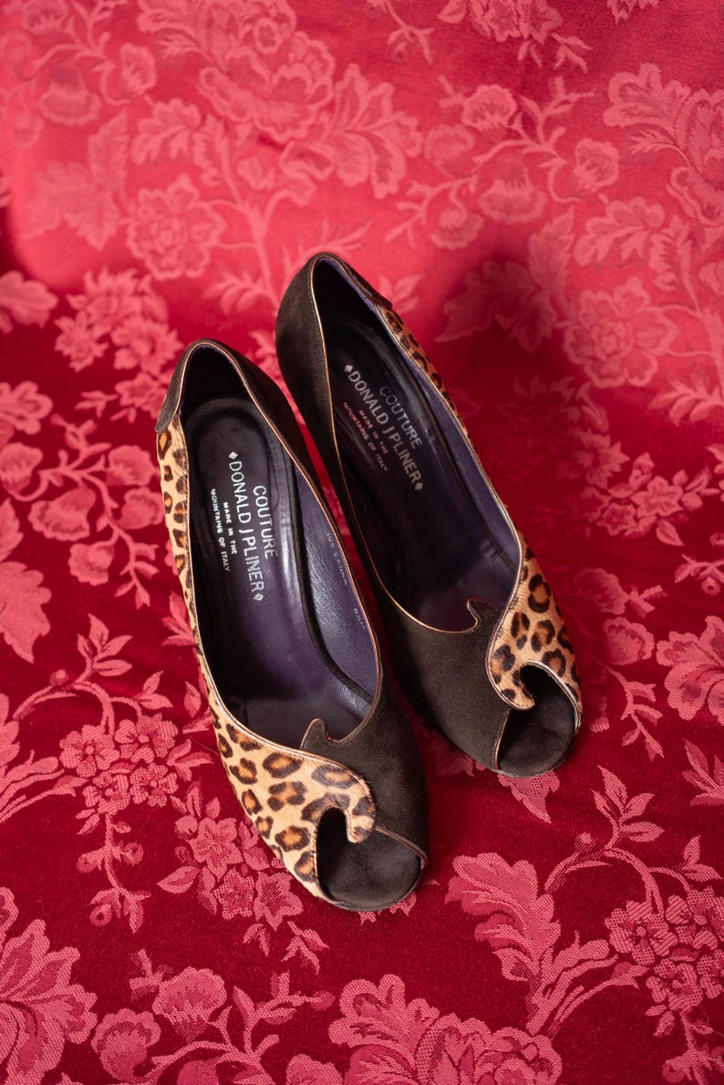 K&ouml;nnte beinhalten: Ein Paar Peep-Toe-Pumps aus dunkelbraunem Wildleder mit Leopardenmuster-Akzenten. Die Innensohlen sind mit dem Text COUTURE DONALD J PLINER MADE IN THE MOUNTAINS OF ITALY versehen. Die Schuhe haben einen hohen Absatz und ein geschwungenes Design.
