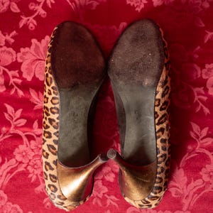 K&ouml;nnte beinhalten: Ein Paar hochhackige Schuhe mit Leopardenmuster auf der Au&szlig;enseite, einem metallisch bronzefarbenen Absatz und einer dunkelbraunen Sohle. Die Gr&ouml;&szlig;e 8.5 M ist deutlich auf der Unterseite jedes Schuhs aufgedruckt und gibt die Passform an.