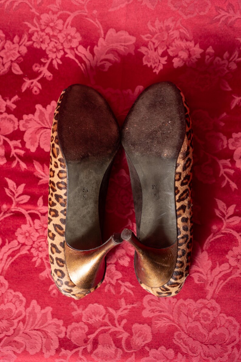 K&ouml;nnte beinhalten: Ein Paar hochhackige Schuhe mit Leopardenmuster auf der Au&szlig;enseite, einem metallisch bronzefarbenen Absatz und einer dunkelbraunen Sohle. Die Gr&ouml;&szlig;e 8.5 M ist deutlich auf der Unterseite jedes Schuhs aufgedruckt und gibt die Passform an.