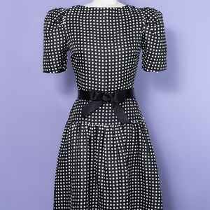 May include: A black dress with a white polka dot pattern, featuring short puff sleeves, a round neckline, and a fitted waist accented by a black satin ribbon belt tied in a bow at the center. The skirt has a drop waist and a flared silhouette.