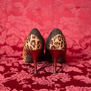 K&ouml;nnte beinhalten: Ein Paar hochhackige Pumps mit einem Obermaterial aus dunkelbraunem Wildleder und einem Leopardenmuster an der Fersenkappe. Die Schuhe haben einen schlanken, dunkel get&ouml;nten Stiletto-Absatz und sind so positioniert, dass die R&uuml;ckseite sichtbar ist.