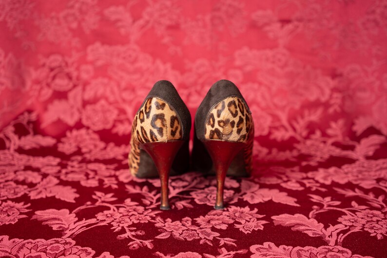 K&ouml;nnte beinhalten: Ein Paar hochhackige Pumps mit einem Obermaterial aus dunkelbraunem Wildleder und einem Leopardenmuster an der Fersenkappe. Die Schuhe haben einen schlanken, dunkel get&ouml;nten Stiletto-Absatz und sind so positioniert, dass die R&uuml;ckseite sichtbar ist.