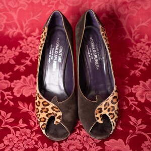 K&ouml;nnte beinhalten: Ein Paar dunkelbraune Wildleder-Pumps mit offener Zehenpartie und Leopardenmuster-Akzenten an den Seiten und vorne. Die dunkelvioletten Innensohlen sind mit dem Text COUTURE DONALD J PLINER MADE IN THE MOUNTAINS OF ITALY bedruckt.