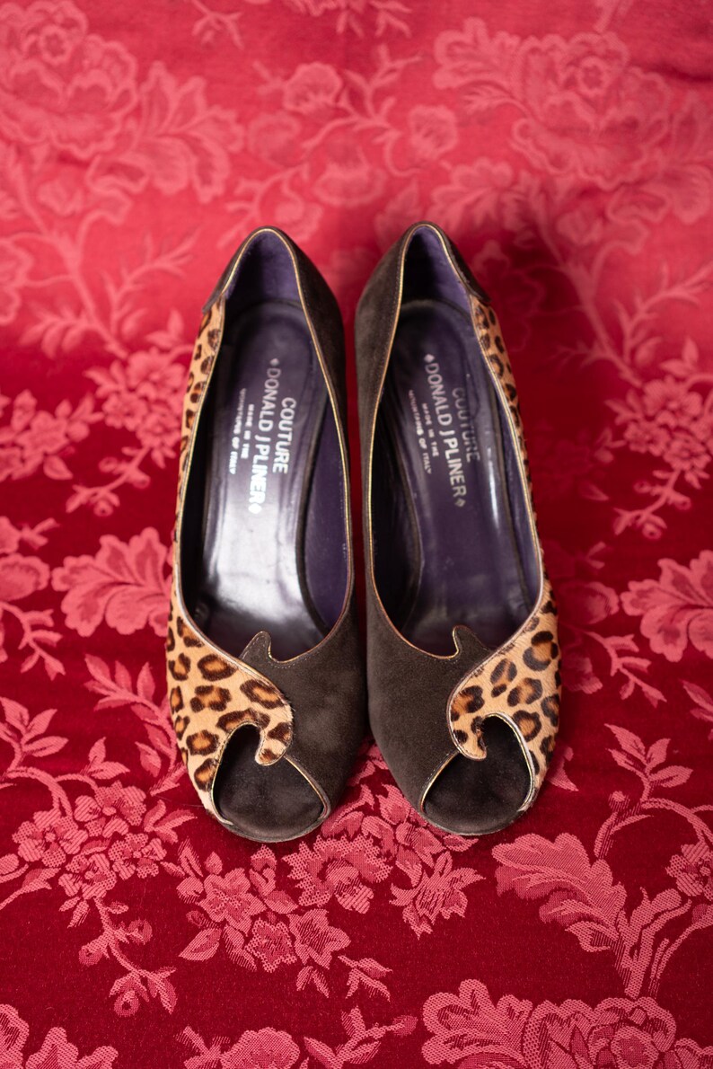 K&ouml;nnte beinhalten: Ein Paar dunkelbraune Wildleder-Pumps mit offener Zehenpartie und Leopardenmuster-Akzenten an den Seiten und vorne. Die dunkelvioletten Innensohlen sind mit dem Text COUTURE DONALD J PLINER MADE IN THE MOUNTAINS OF ITALY bedruckt.