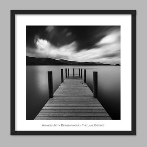 May include: A black and white photograph of a wooden jetty extending out into a still lake. The sky is cloudy and the mountains in the distance are barely visible. The text "Ashness Jetty Derwentwater - The Lake District" is at the bottom of the image.