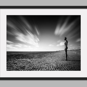 May include: A black and white photograph of a solitary figure standing on a beach, gazing out at the horizon. The sky is filled with dramatic, swirling clouds.