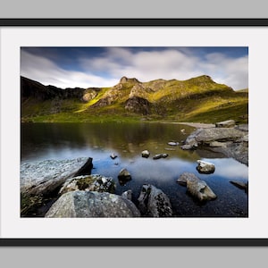 May include: A scenic landscape photograph of a lake reflecting the sky and mountains in the distance. The lake is surrounded by rocks and green hills. The sky is a light blue with white clouds.