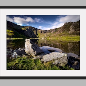 May include: A framed landscape photograph of a mountain lake. The image shows a tranquil lake reflecting the mountains and sky. Large rocks and green grass are in the foreground, with a blue sky and clouds above.