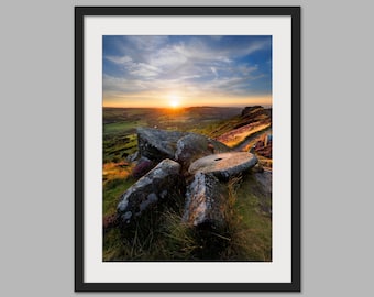 May include: A scenic sunset over a green valley with large rocks in the foreground. The sky is a vibrant blue with fluffy clouds and the sun is setting behind the horizon. The rocks are grey and weathered, and the grass is green and lush.