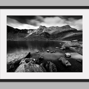May include: A black and white photograph of a mountain lake with a rocky shoreline. The water is calm and reflects the sky and clouds. The mountains in the background are covered in trees and snow.