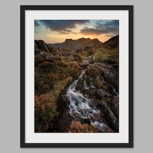 May include: A scenic photograph of a waterfall cascading down rocky terrain in a mountainous landscape. The sun is setting behind the mountains, casting a warm glow on the scene. The water is flowing smoothly, creating a sense of tranquility.
