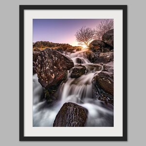 May include: A long exposure photograph of a waterfall cascading over rocks in a mountainous landscape. The water is blurred, creating a silky effect. The sky is a vibrant purple and orange, with a lone tree silhouetted against the sunset.