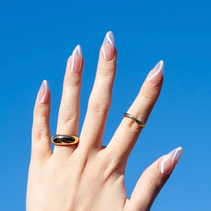 Puede incluir: Una mano con u&ntilde;as largas y almendradas pintadas con un dise&ntilde;o de punta blanca. Las u&ntilde;as est&aacute;n pintadas de un color rosa claro. La mano lleva dos anillos de oro.